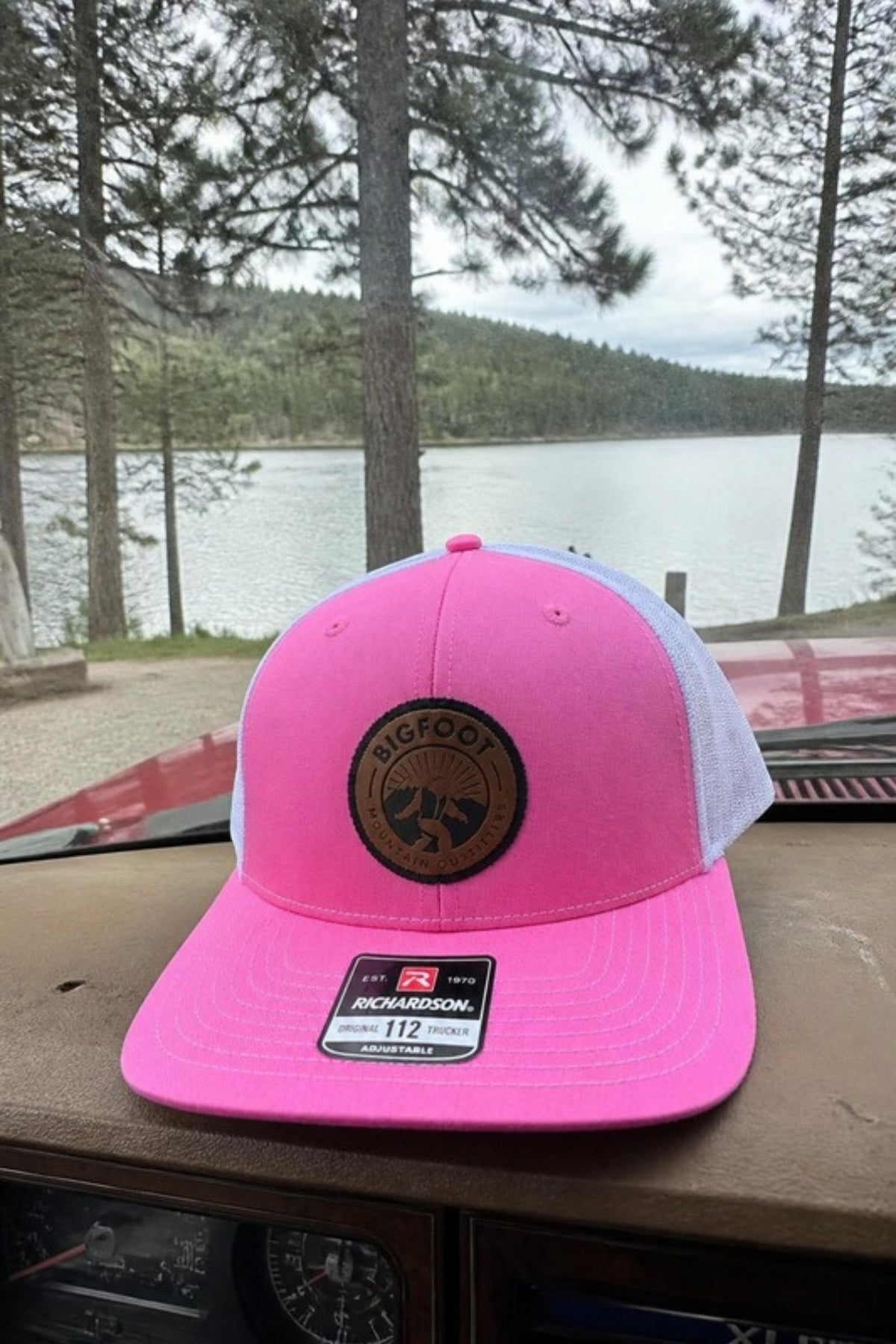 Bigfoot Mountain Outfitters Original Patch Logo mesh back hat in Pink/White sitting on a pickup dashboard by a mountain lake, a fun trucker cap for road trips, lake days, camping and everyday wear