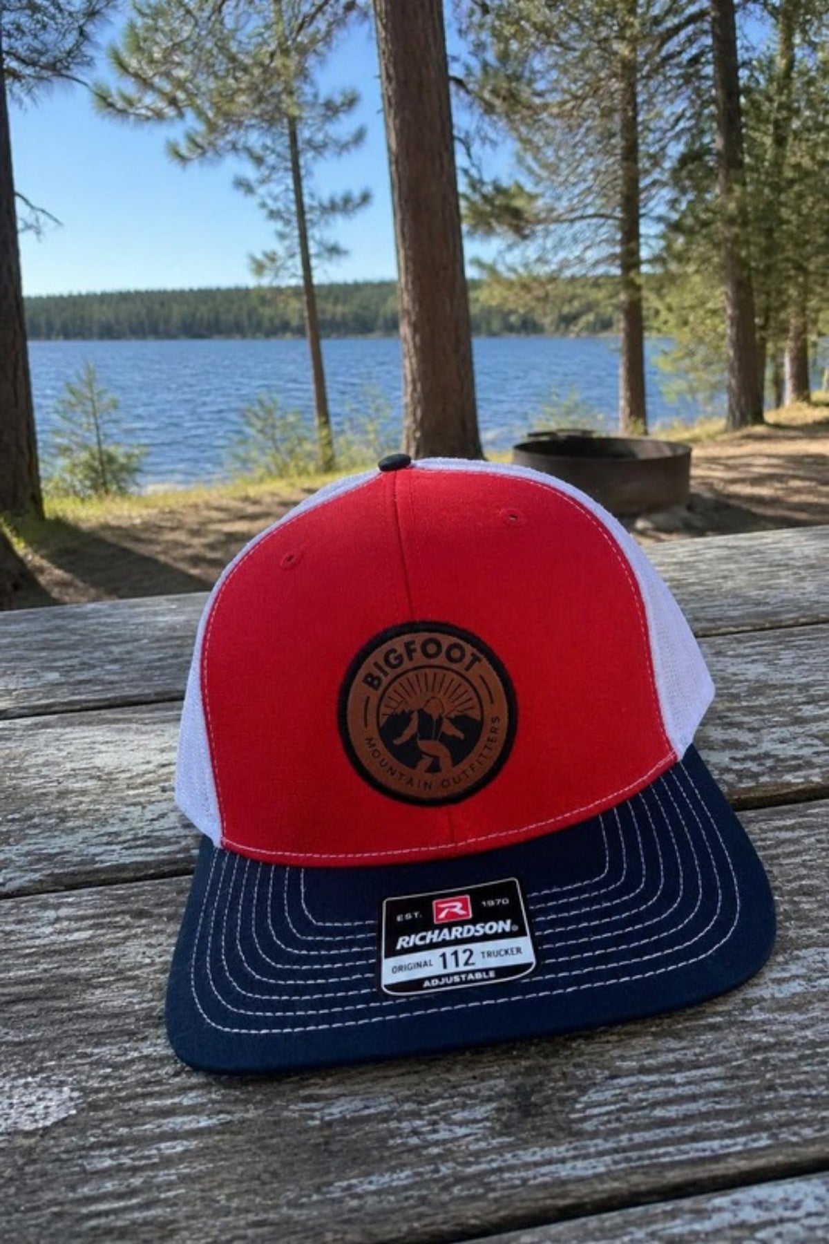 Bigfoot Mountain Outfitters Original Patch Logo mesh back hat in Red/Navy/White on a picnic table at a lakeside campground, a classic trucker cap for camping, fishing, road trips and Fourth of July weekends
