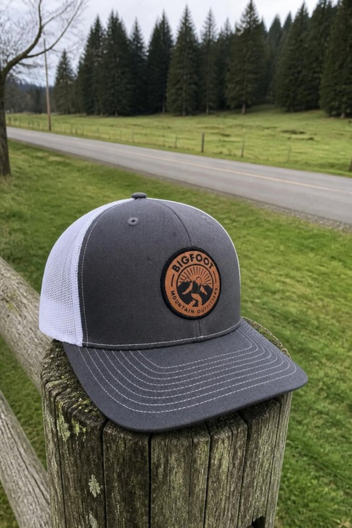 Bigfoot Mountain Outfitters BMO Mesh Hat in Grey/White on a country fence post, a leather-patch trucker hat for back roads, yard work, fishing and everyday wear