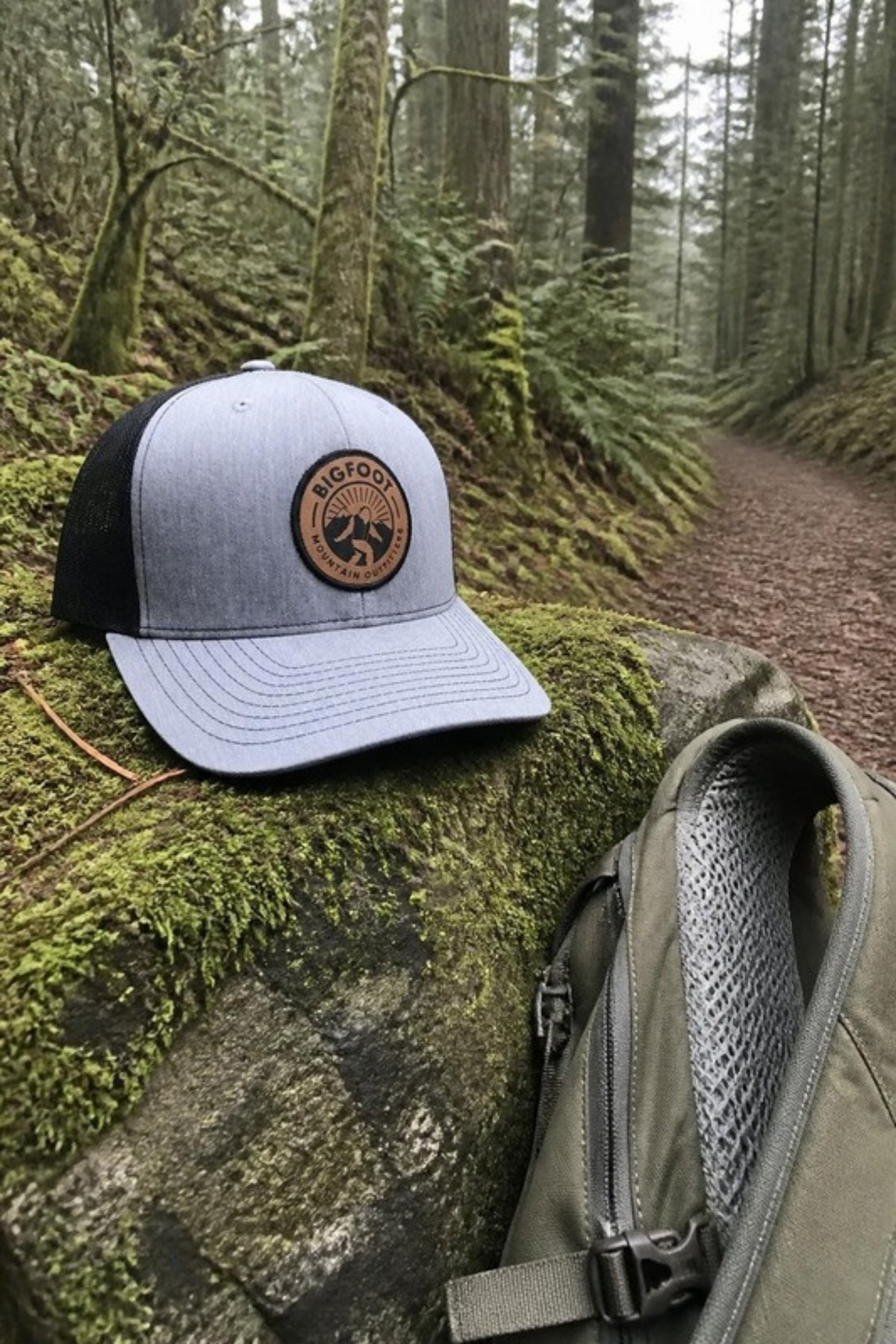 Bigfoot Mountain Outfitters BMO Mesh Hat in Heather Grey/Black on a mossy boulder on a forest trail, a leather-patch trucker hat for hikes, camping and everyday Pacific Northwest wear