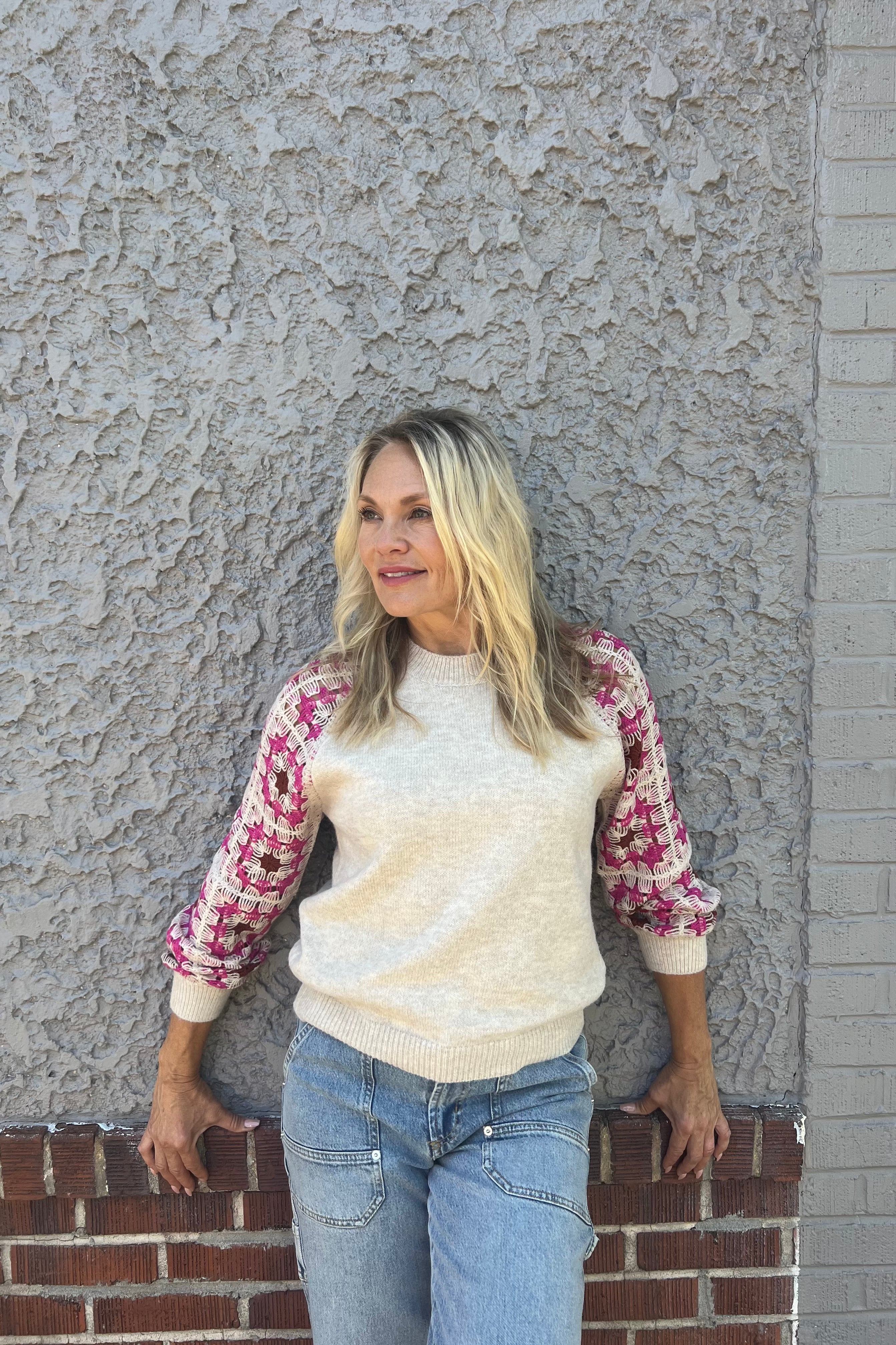 The Amelia Crochet Sweater features an oat-colored body contrasted by charming pink and brown detailed sleeves with cuffs. It includes a classic crew neckline and is crafted to be incredibly soft and lightweight for comfortable wear.