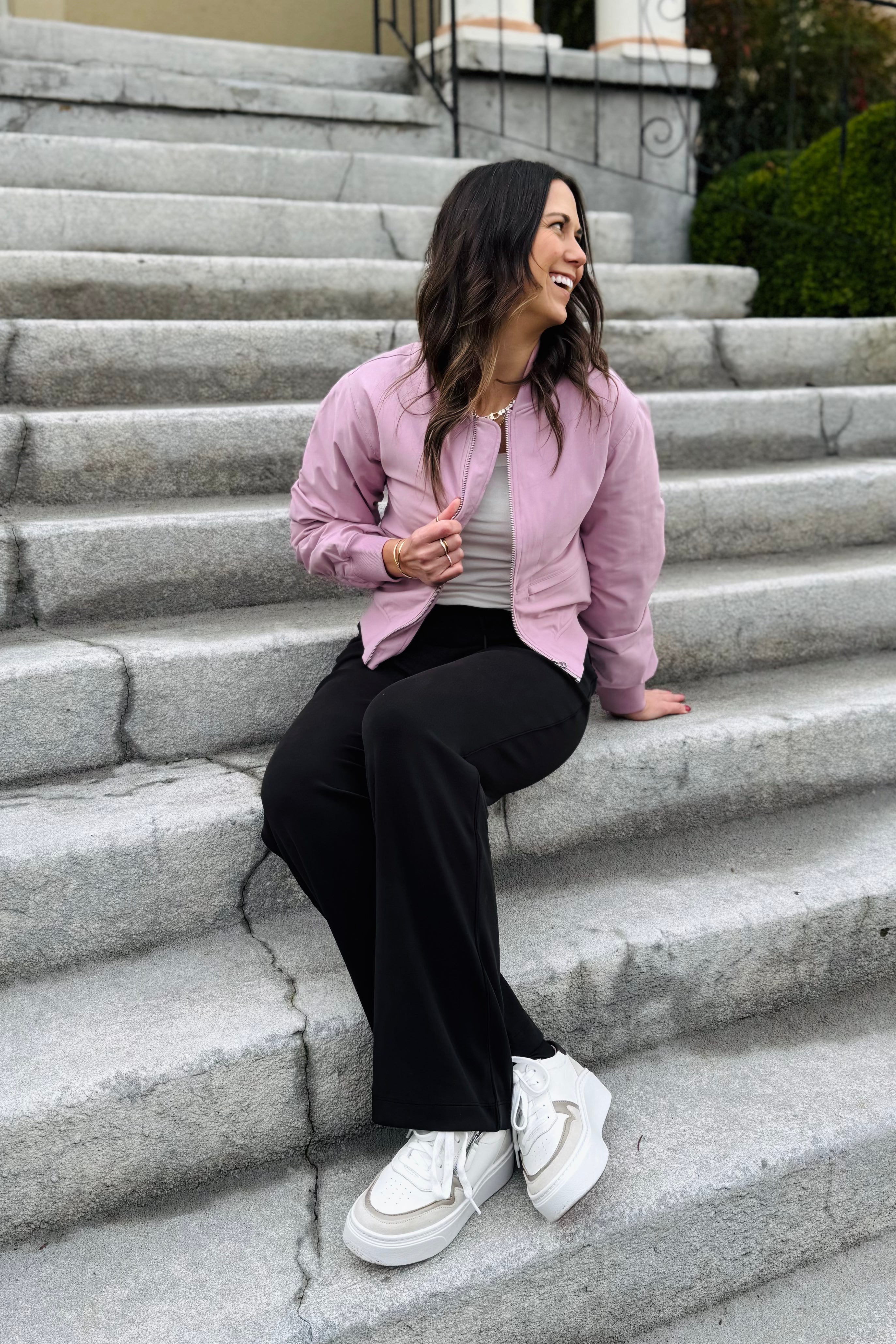 Woman sitting on stone steps wearing a pink jacket, black pants, and white sneakers.