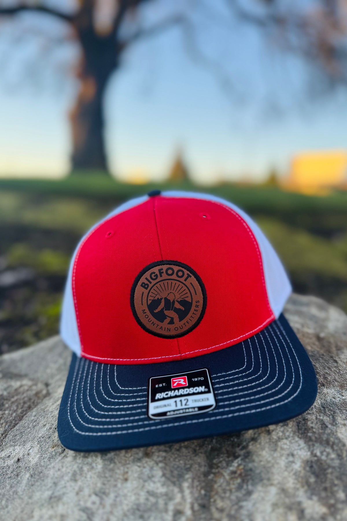Bigfoot Mountain Outfitters Original Patch Logo mesh back hat in Red/Navy/White on a picnic table at a lakeside campground, a classic trucker cap for camping, fishing, road trips and Fourth of July weekends