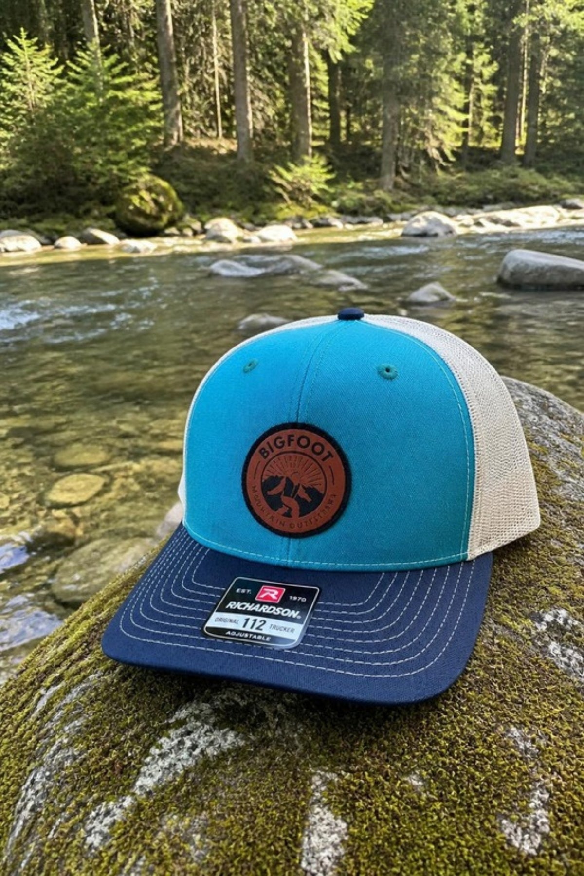 Bigfoot Mountain Outfitters Original Patch Logo mesh back hat in Teal/Navy/Cream on a mossy boulder beside a mountain stream, a breathable trucker cap for fly fishing, hiking, river days and everyday wear
