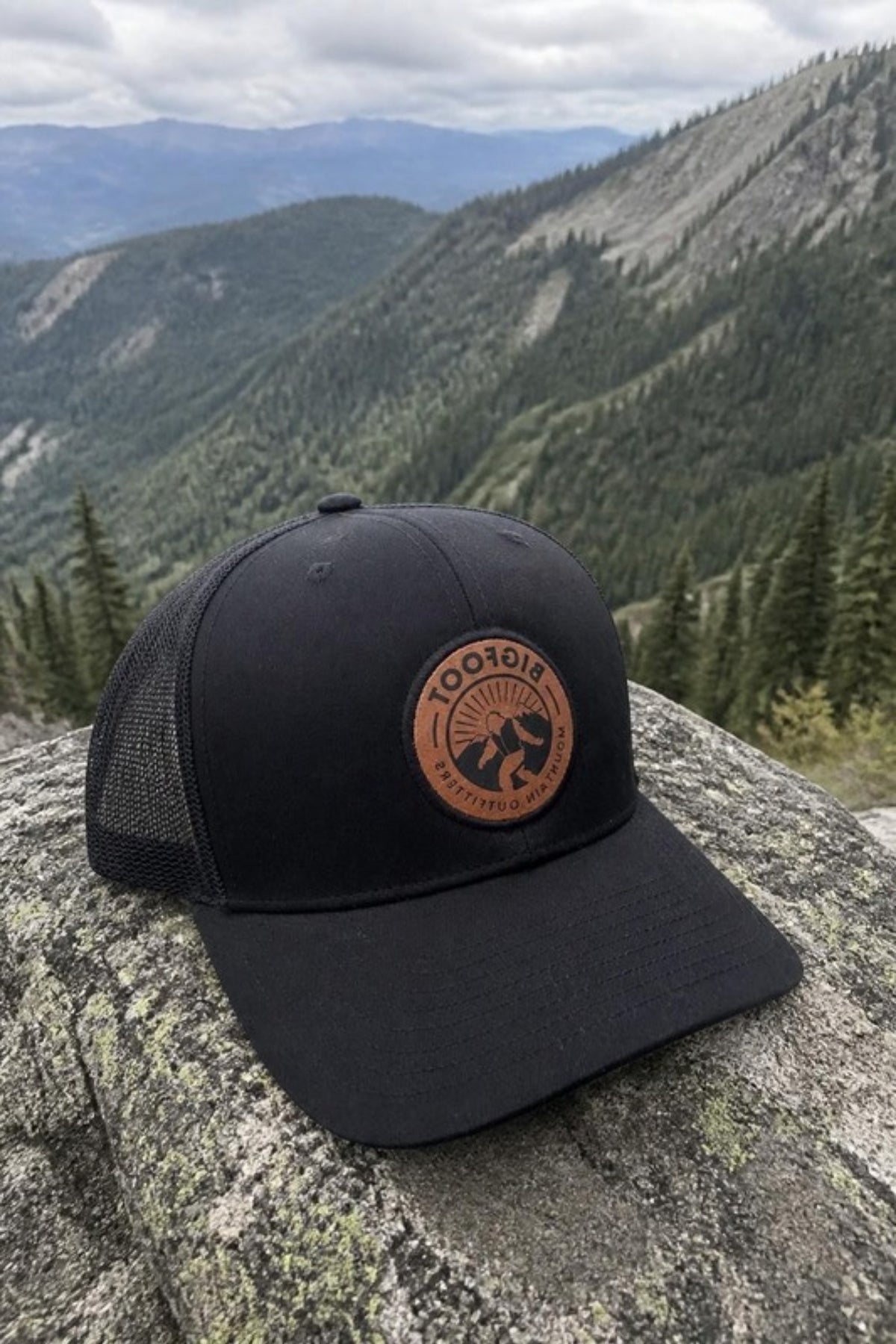 Bigfoot Mountain Outfitters BMO Mesh Hat in Black on a mountain overlook rock, an all-black leather-patch trucker hat for hikes, road trips and everyday wear