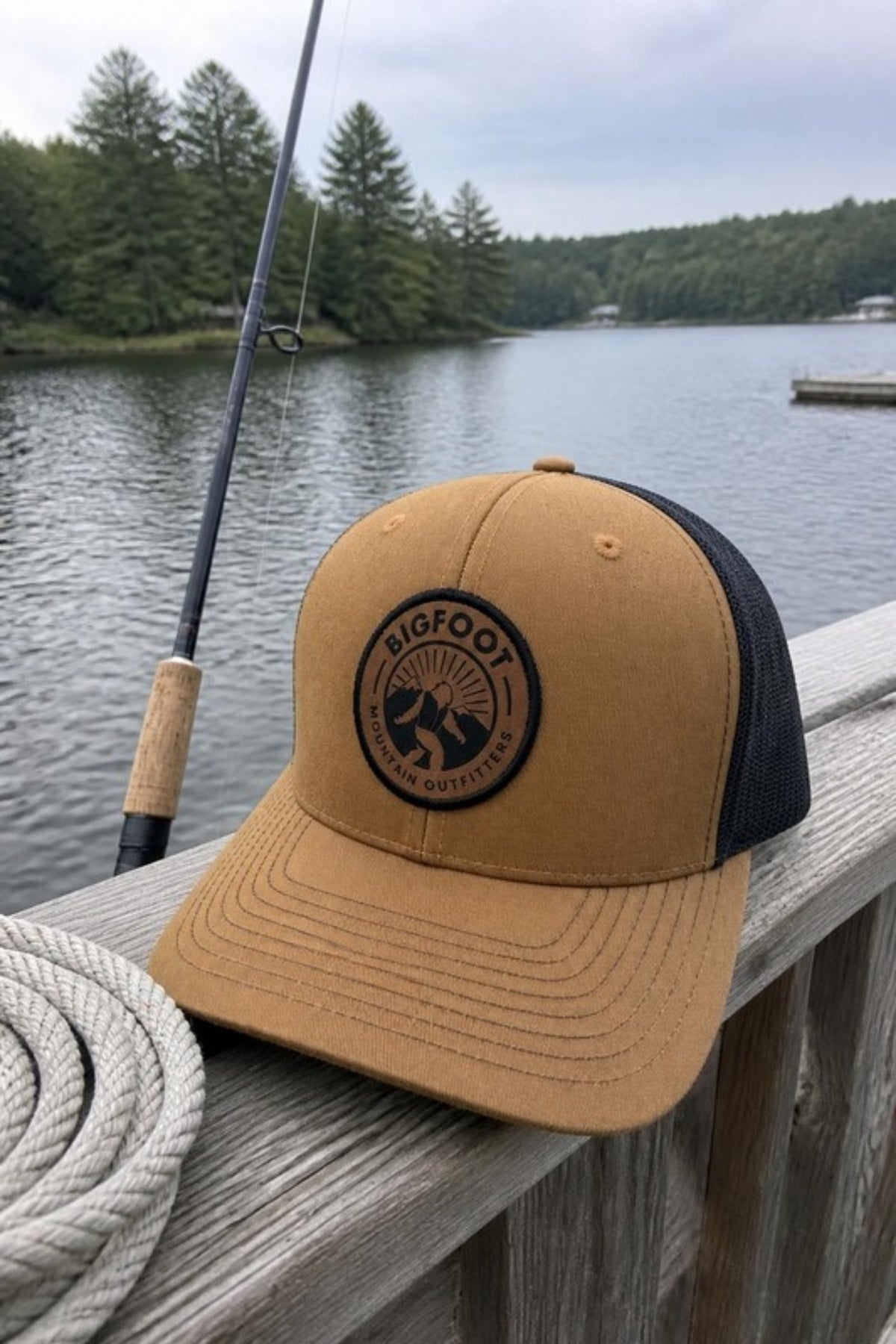 Bigfoot Mountain Outfitters BMO Mesh Hat in Caramel/Black on a lake dock rail by a fishing rod, a leather-patch trucker hat for lake days, fishing and summer weekends