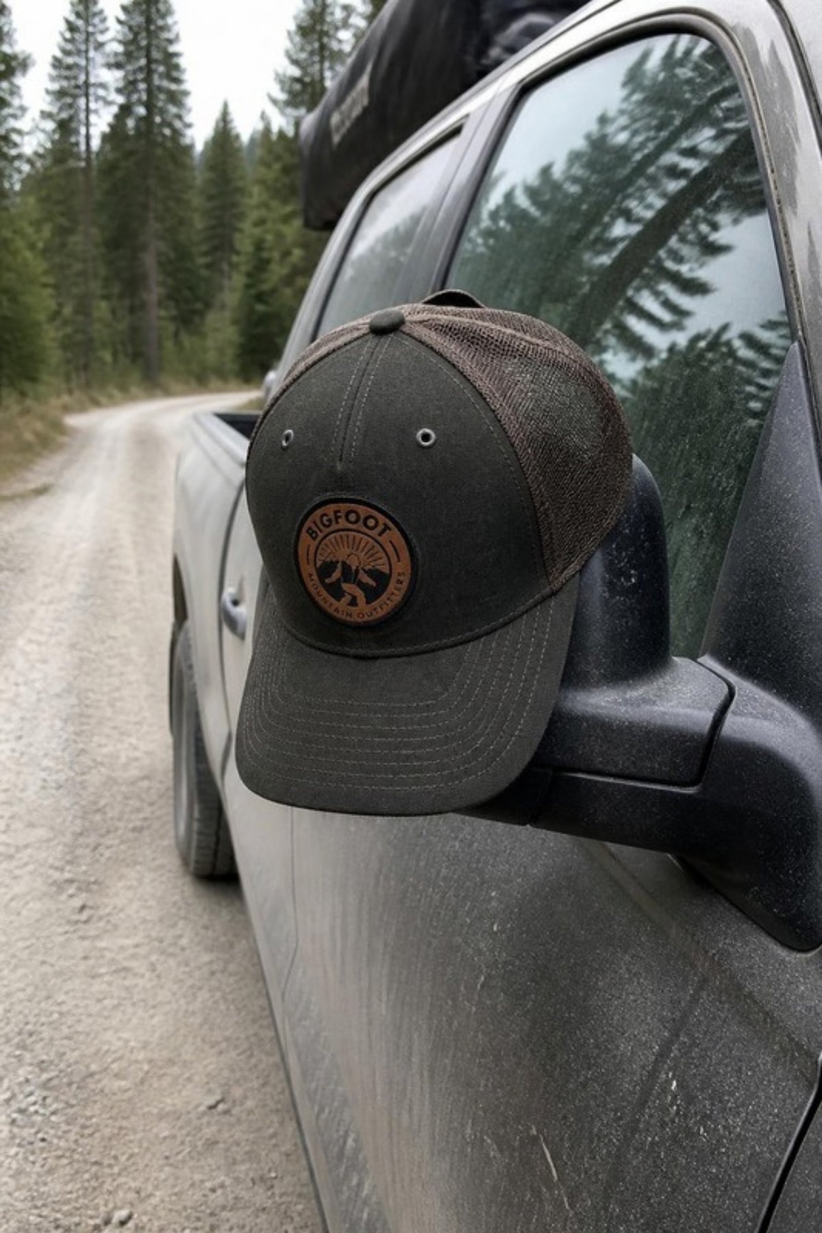 Bigfoot Mountain Outfitters BMO Mesh Hat in Dark Olive/Coffee hanging on a truck mirror on a forest road, a leather-patch trucker hat for back roads, camping, fishing and everyday wear