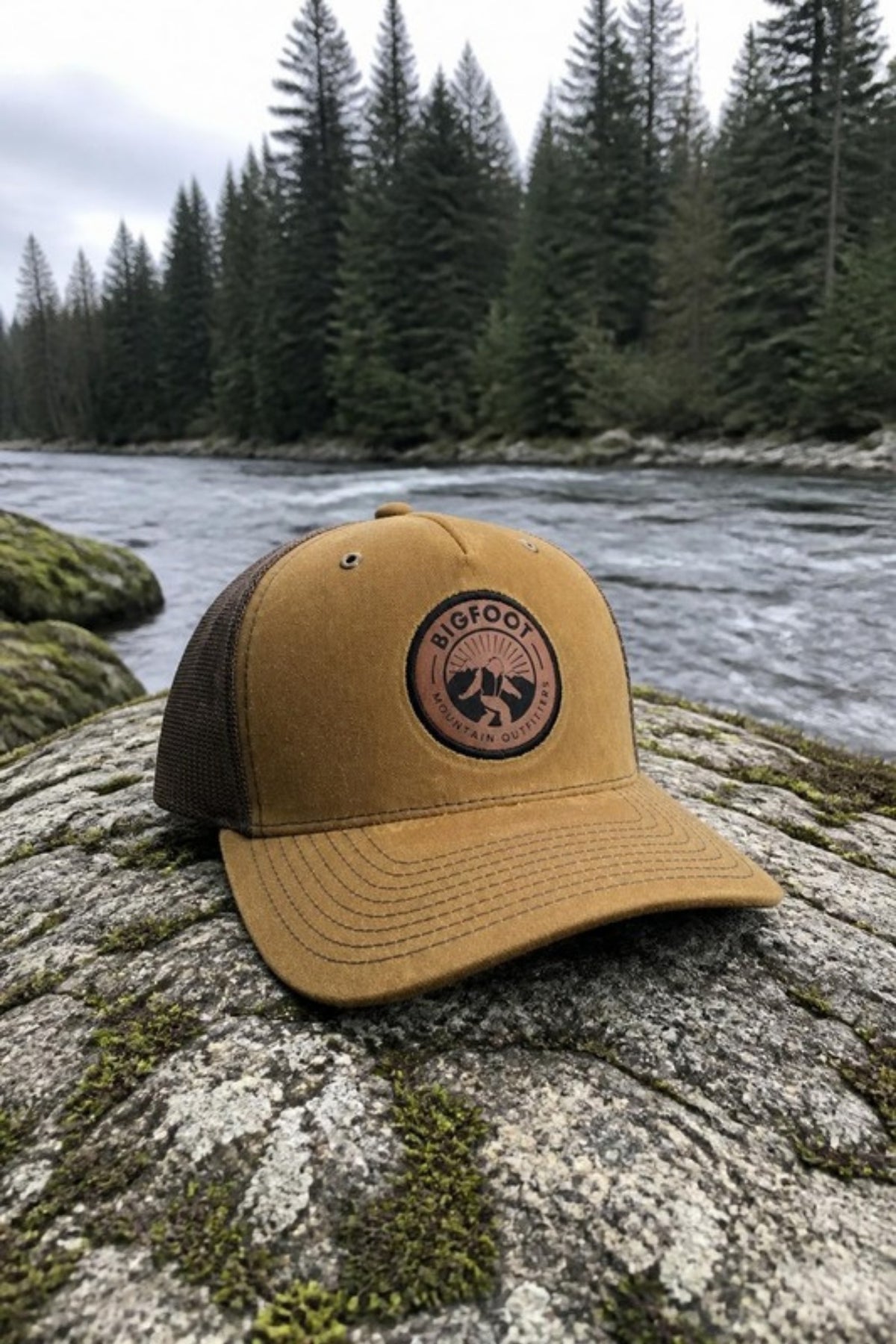 Bigfoot Mountain Outfitters BMO Mesh Hat in Whiskey/Brown on a mossy river rock, a classic leather-patch trucker hat for fishing, hiking, road trips and everyday Pacific Northwest adventures