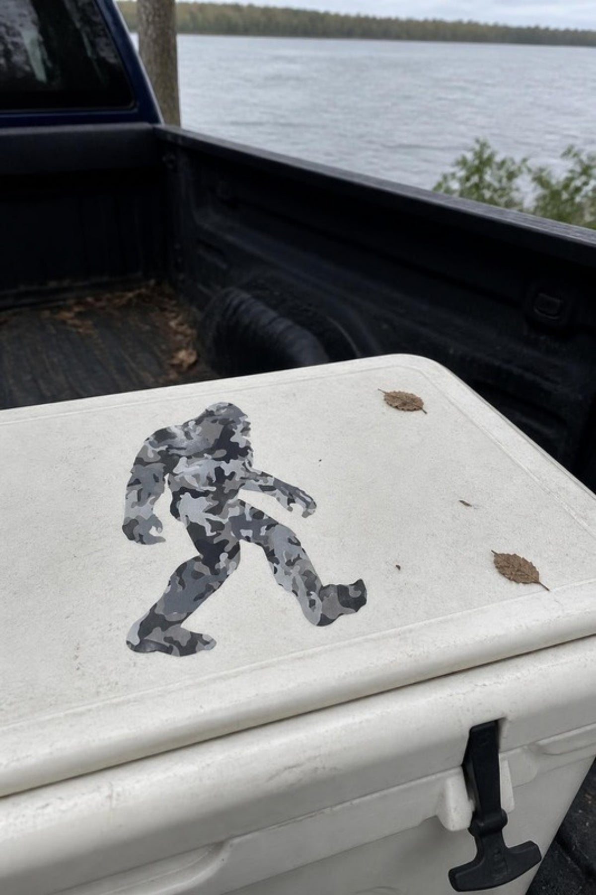 Paradise Cay Publications Camo Sasquatch sticker in grey camo on a cooler lid in a truck bed, a laminated vinyl Bigfoot decal for coolers, trucks, water bottles and laptops