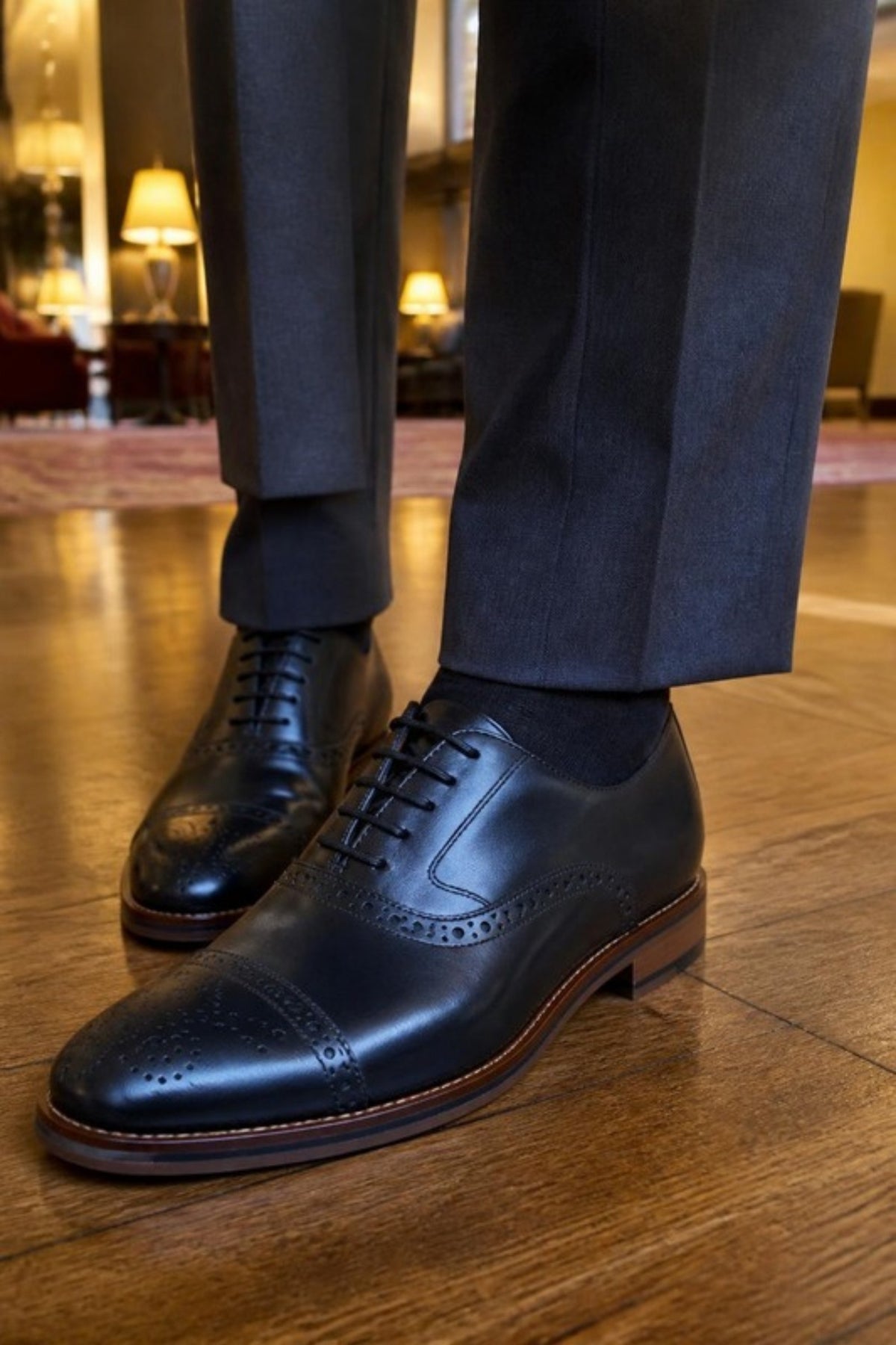 Johnston & Murphy Conard 2.0 cap toe oxford in Black full grain leather with brogue detailing, worn with charcoal suit trousers in a hotel lobby, a cushioned, temperature-regulating dress shoe for the office, weddings, formal events and all-day wear