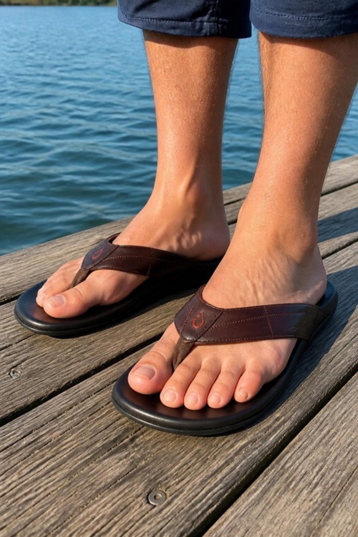 OluKai Hokulea Kia leather sandal in Dark Brown full-grain leather with a molded cushioned footbed, worn on a lakeside dock in summer, a premium men's flip-flop for beach days, boating, vacations and relaxed weekend errands