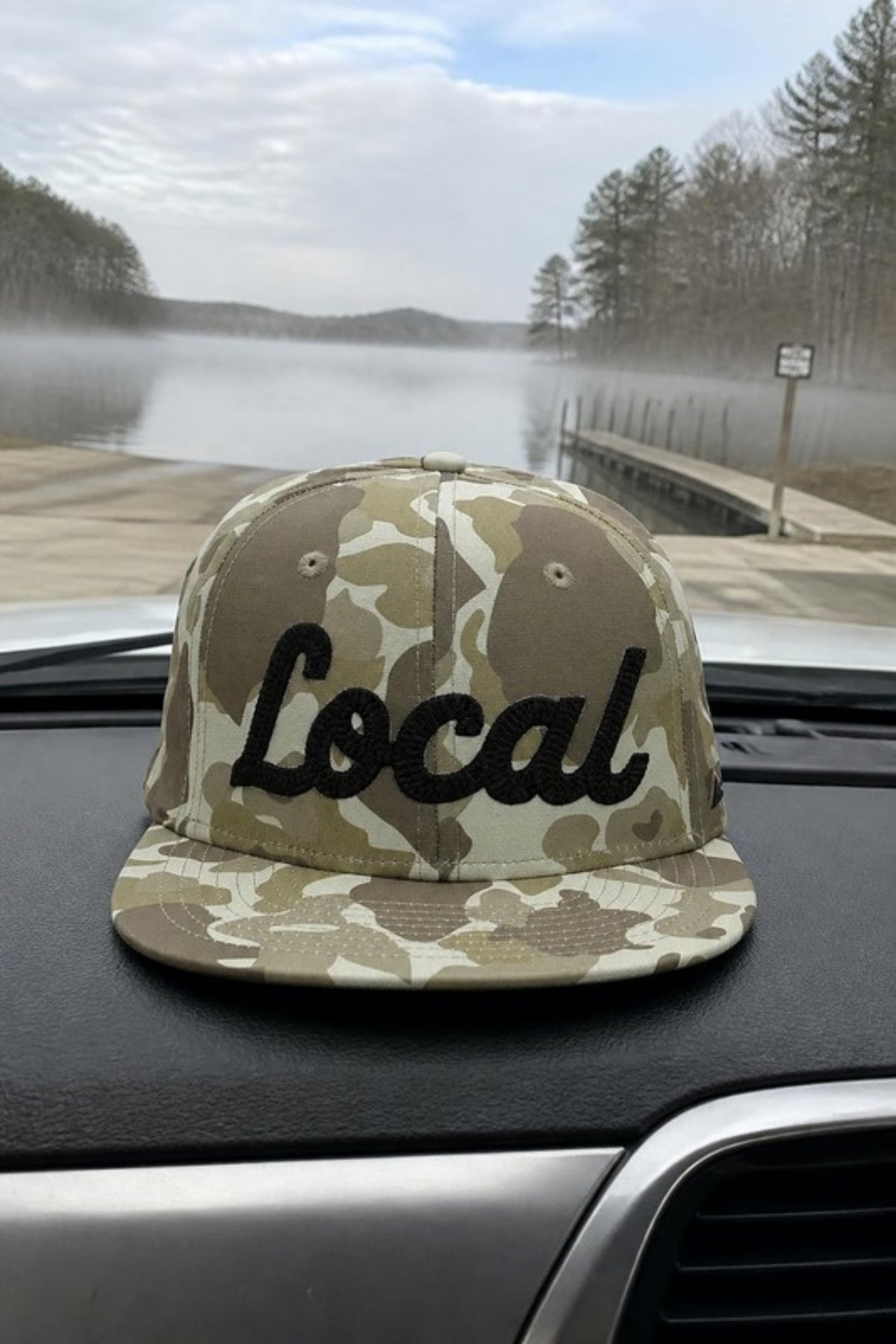 Paramount Outdoors Local Camo Hat on a truck dashboard at the boat launch, a duck camo flat-brim snapback with Local script embroidery for fishing trips, hunting season and hometown pride