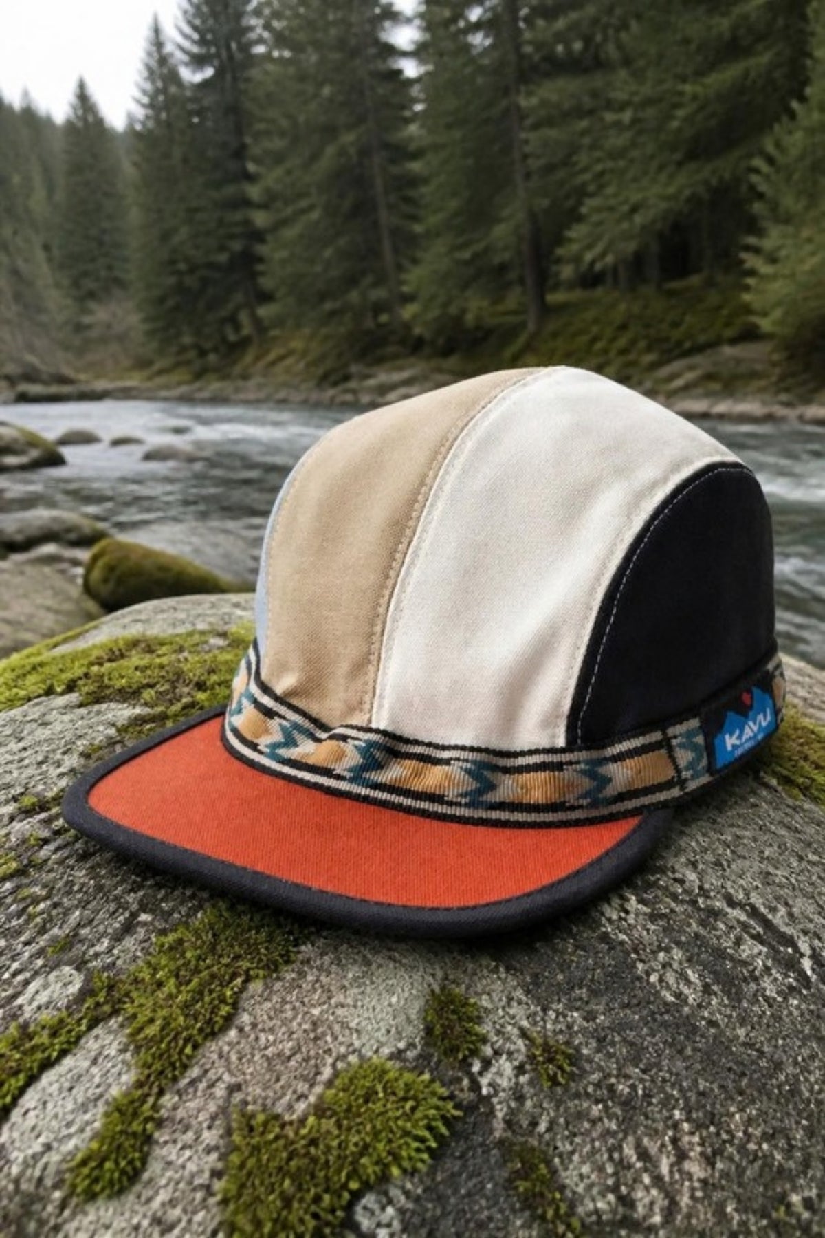 KAVU Organic Strapcap in tan, cream, black and blue with a burnt-orange brim and woven strap, resting on a mossy boulder beside a mountain river. A packable organic-cotton camp cap made for hiking, fishing trips and riverside days.